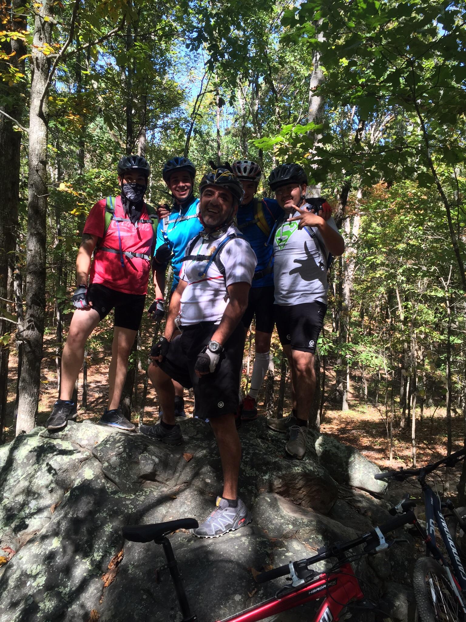 A group of five cyclists posing together on a rock in a wooded area. They are wearing helmets and various cycling attire, with some bicycles parked nearby. The background features lush green trees and a clear blue sky, indicating a sunny day. Oak Mountain State Park mountain bike trail.