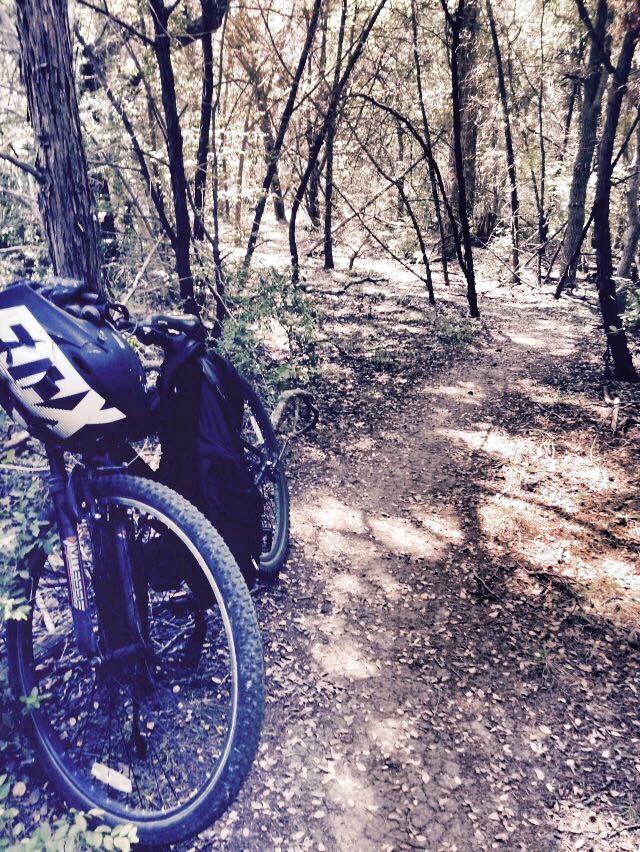 Hyper Havoc: A mountain bike resting on a trail surrounded by trees, with dappled sunlight filtering through the foliage. The path is dirt and runs through a woodland area.