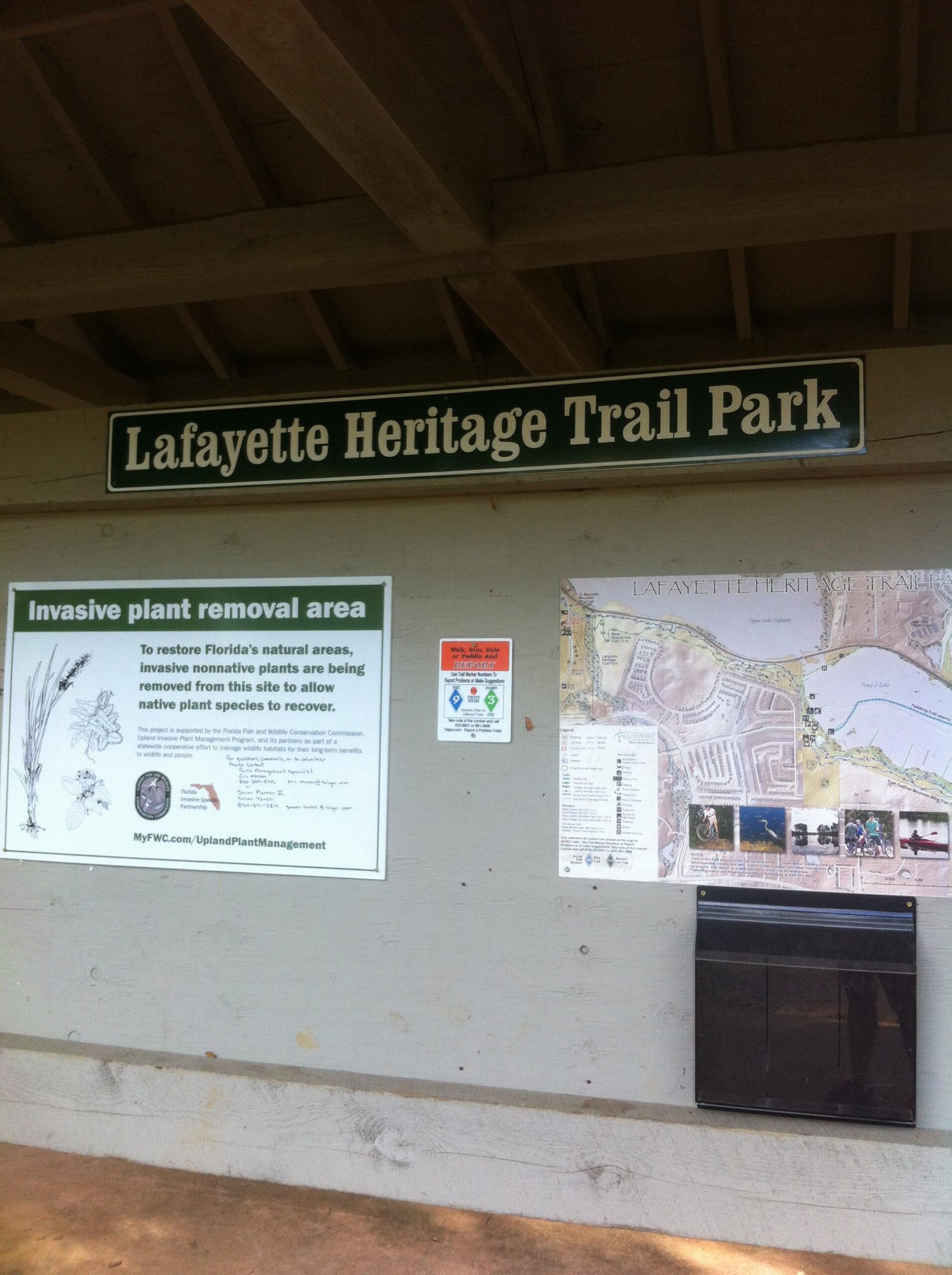 Signage at Lafayette Heritage Trail Park, featuring information about invasive plant removal efforts and a map of the park. The sign explains the initiative to restore native plants in the area, encouraging visitors to support conservation efforts. Various informational posters and a drop box for materials are also visible. Cadillac mountain bike trail.