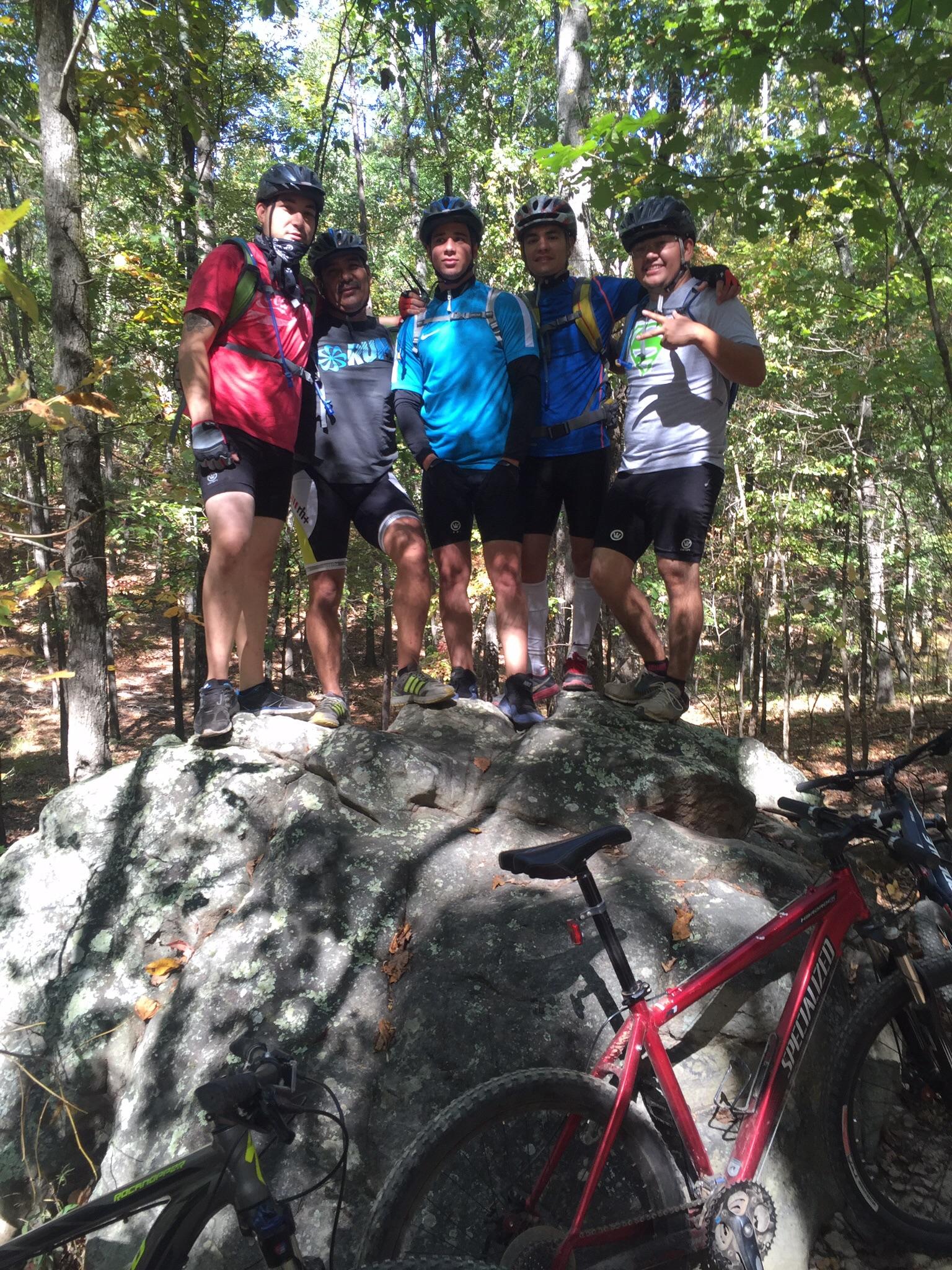 Group of five mountain bikers posing together on a large rock in a forested area, with bicycles nearby. The scene captures the vibrant autumn colors of the trees and shows the cyclists wearing helmets and biking gear. Oak Mountain State Park Bump Trail mountain bike trail.