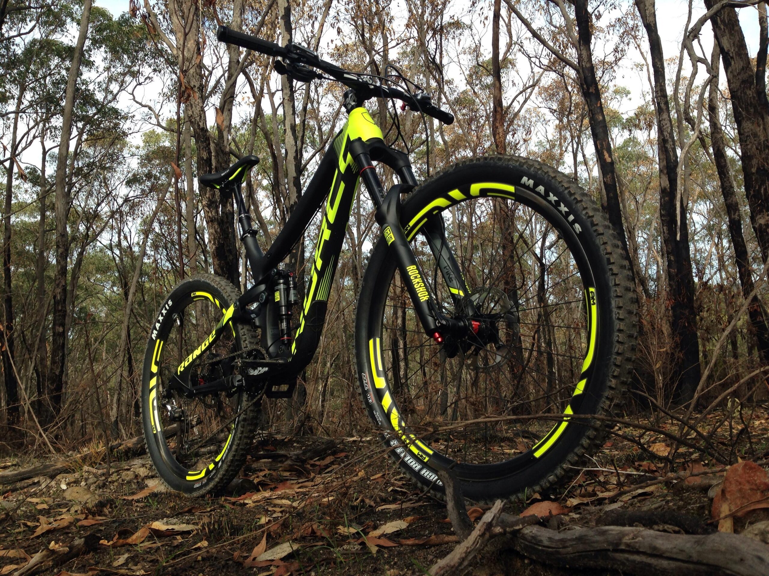 Norco Range Carbon: A mountain bike with a black and neon yellow frame is positioned on a forest floor surrounded by trees and scattered leaves. The bike features thick tires with the brand name "Maxxis" visible, and the setting suggests an outdoor trail or rugged terrain.
