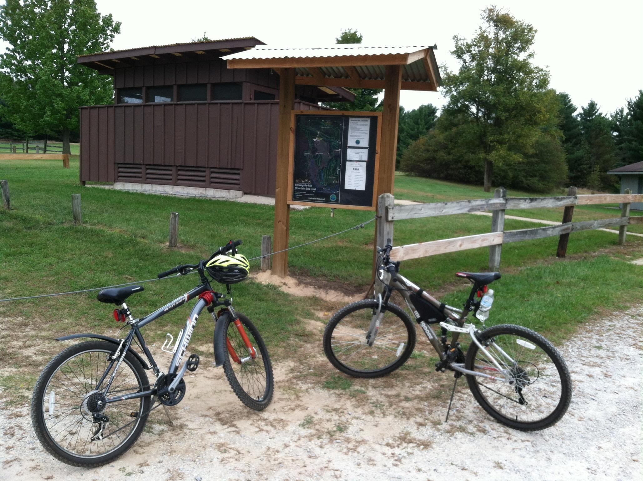 Mongoose Mongoose Ledge 2.1: Two bicycles are parked in front of a trail map sign at a recreational area. A wooden building is visible in the background, surrounded by green grass and trees. The scene conveys a peaceful outdoor setting for biking or hiking.