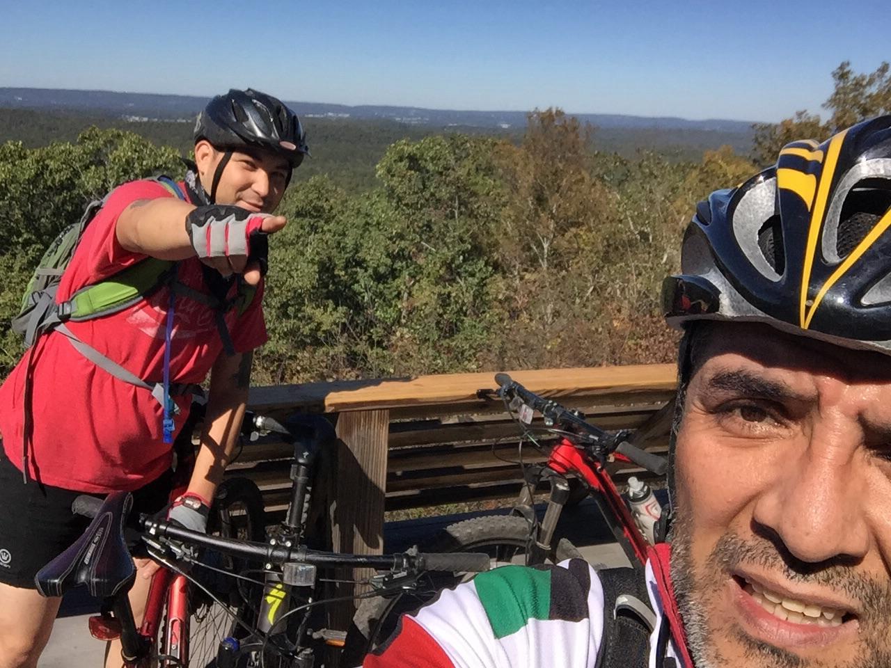 Two mountain bikers pose for a selfie against a scenic backdrop of rolling hills and blue skies. One biker is playfully pointing toward the camera while wearing a red shirt and a backpack, and the other, wearing a helmet and a multicolored jersey, smiles at the camera. Both bikes are parked beside them. Oak Mountain State Park Bump Trail mountain bike trail.