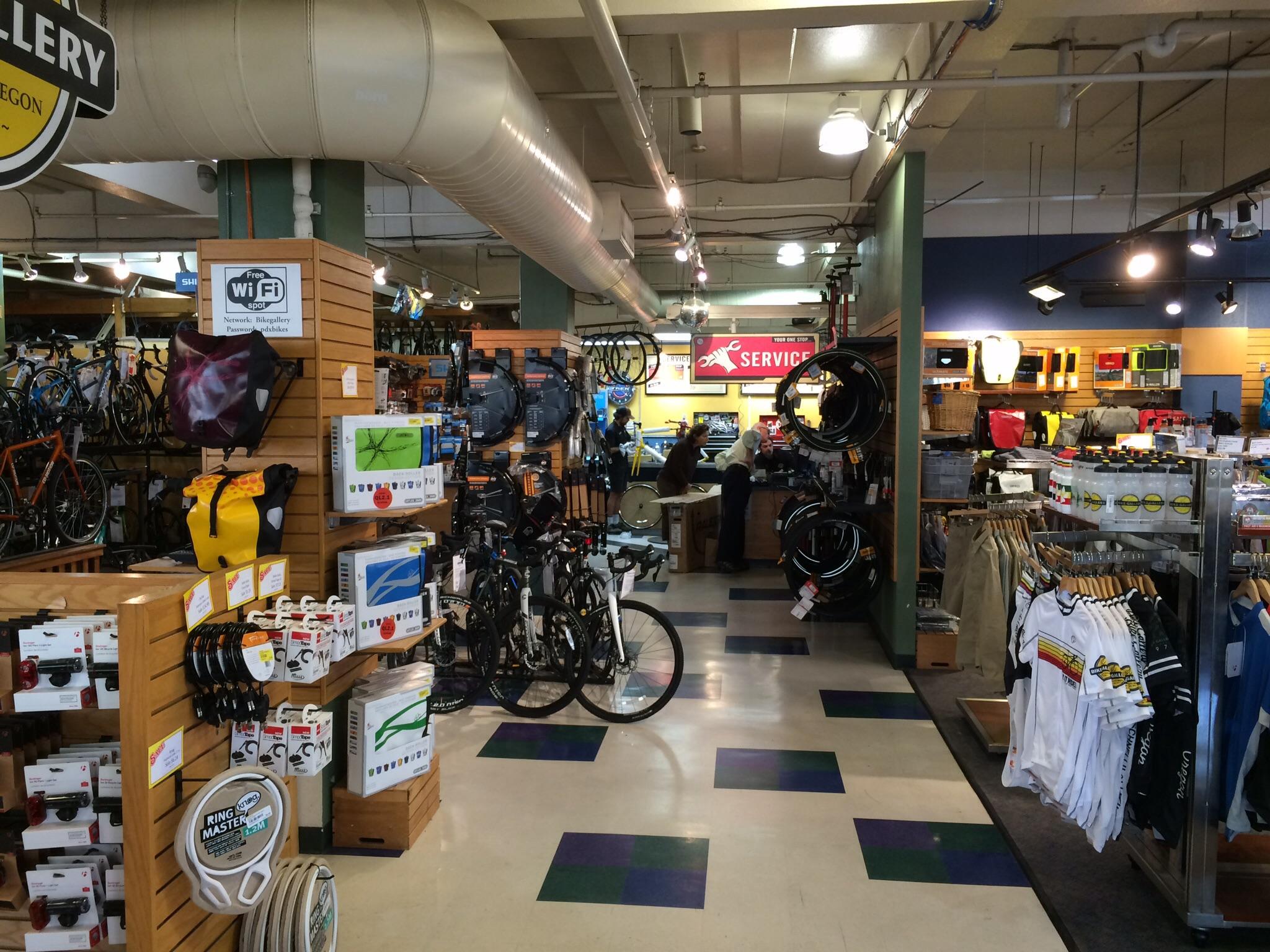 A view of the interior of a bicycle shop featuring a variety of bicycles, biking accessories, and apparel. The store displays multiple products on wooden shelving, including bags, lights, and gear. At the back, a service area can be seen with staff assisting customers. The space is well-lit with a colorful tiled floor.