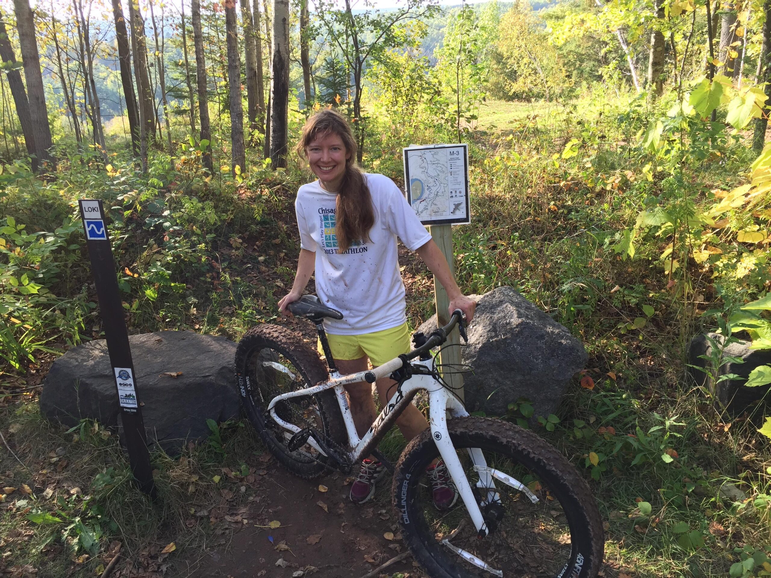Salsa Beargrease: A smiling person stands next to a mountain bike at a trail junction in a forested area. The individual is wearing a white shirt and yellow shorts, with long hair cascading over one shoulder. In the background, there are trees with green leaves and a trail sign labeled "LOKI." A map sign can also be seen, indicating the nearby biking trails. The ground is dirt and slightly muddy, suggesting recent use of the trail.