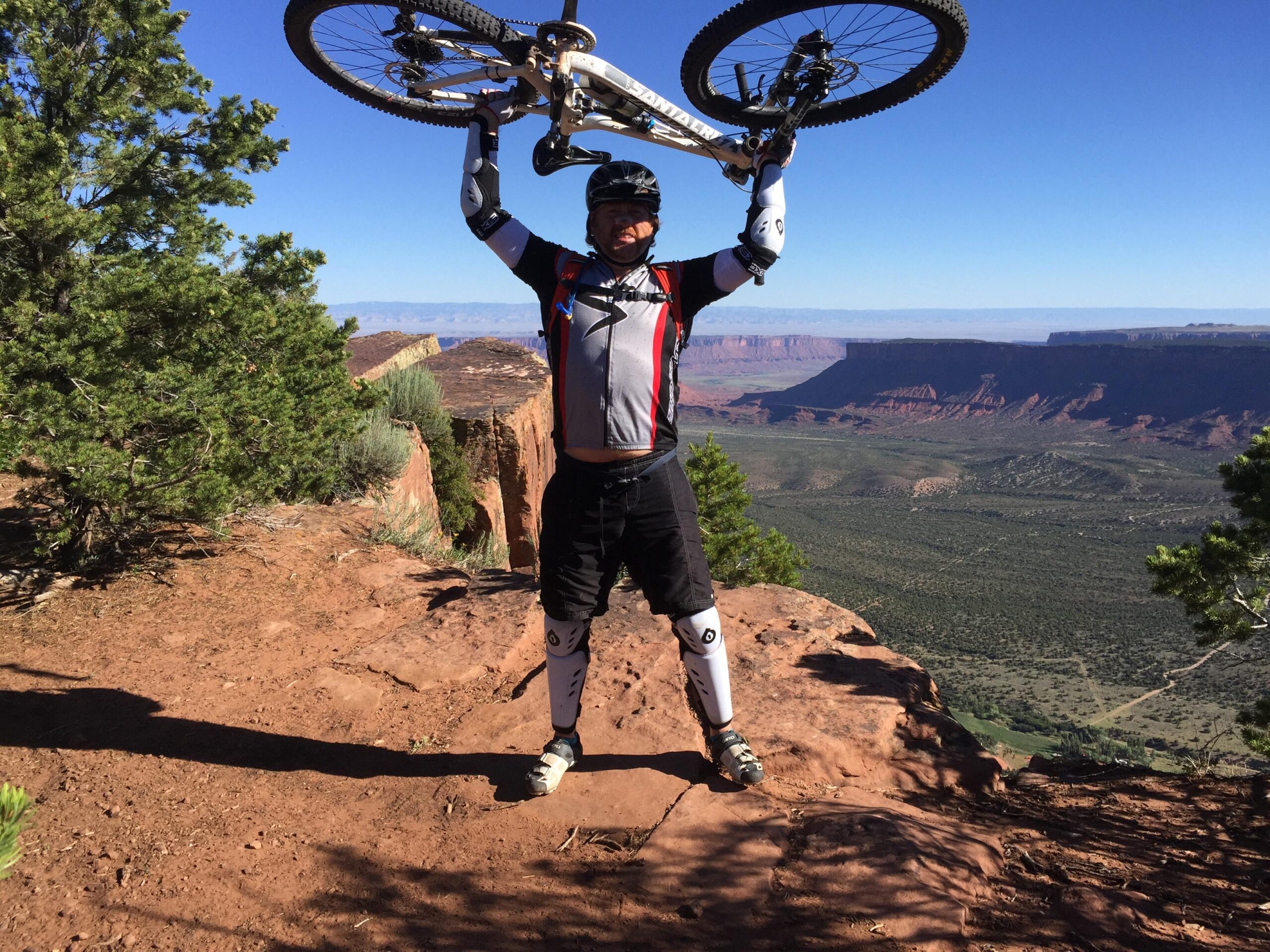 A mountain biker triumphantly raises their bike above their head while standing on a rocky ledge, with a beautiful, expansive landscape of red rock formations and green vegetation in the background. The sky is clear and blue, adding to the sense of achievement and adventure. The Whole Enchilada mountain bike trail.