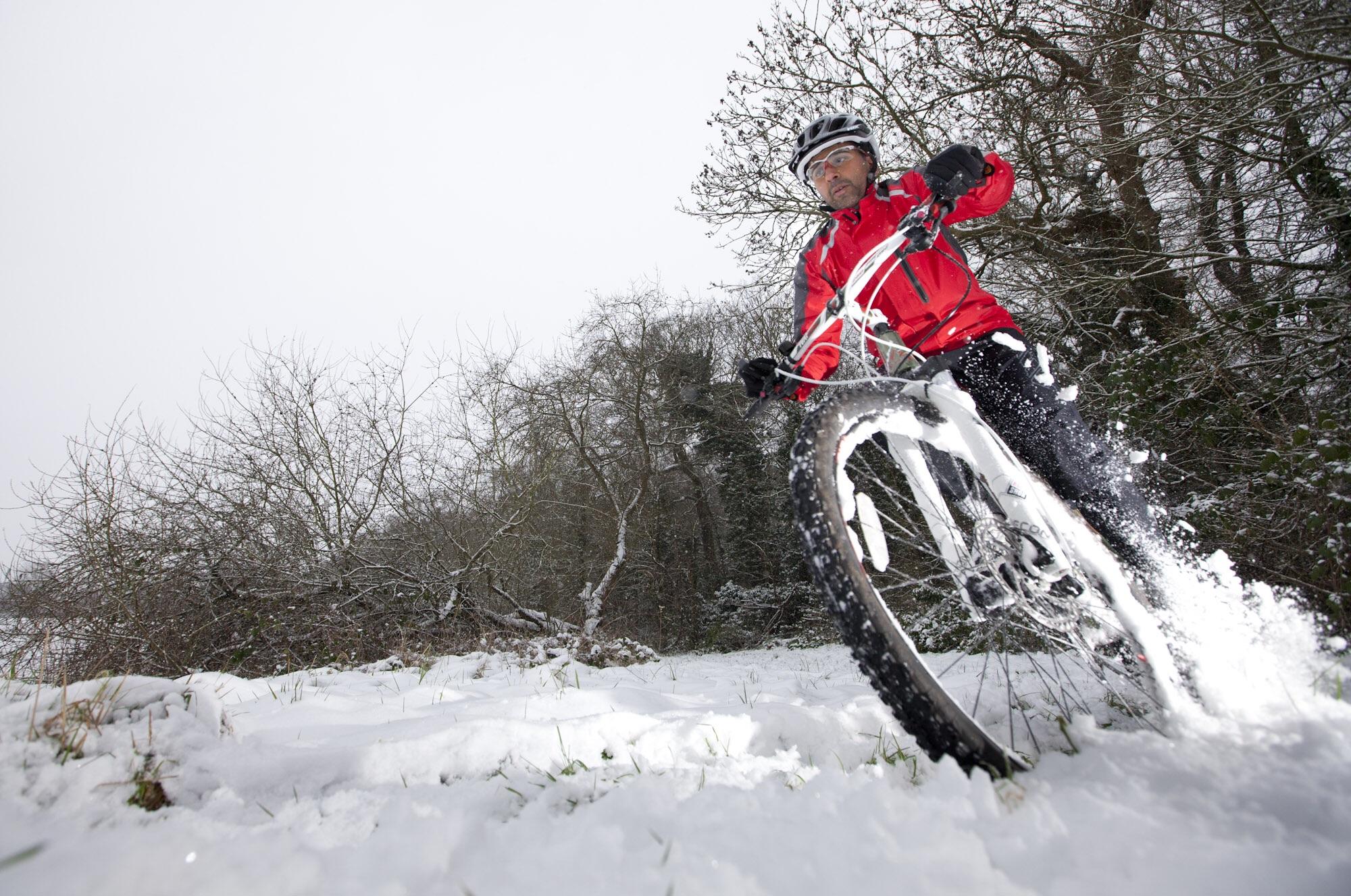 A cyclist in a bright red jacket riding a mountain bike through a snowy landscape, kicking up snow as they navigate a winter trail surrounded by leafless trees. Gabrielino Trail mountain bike trail.