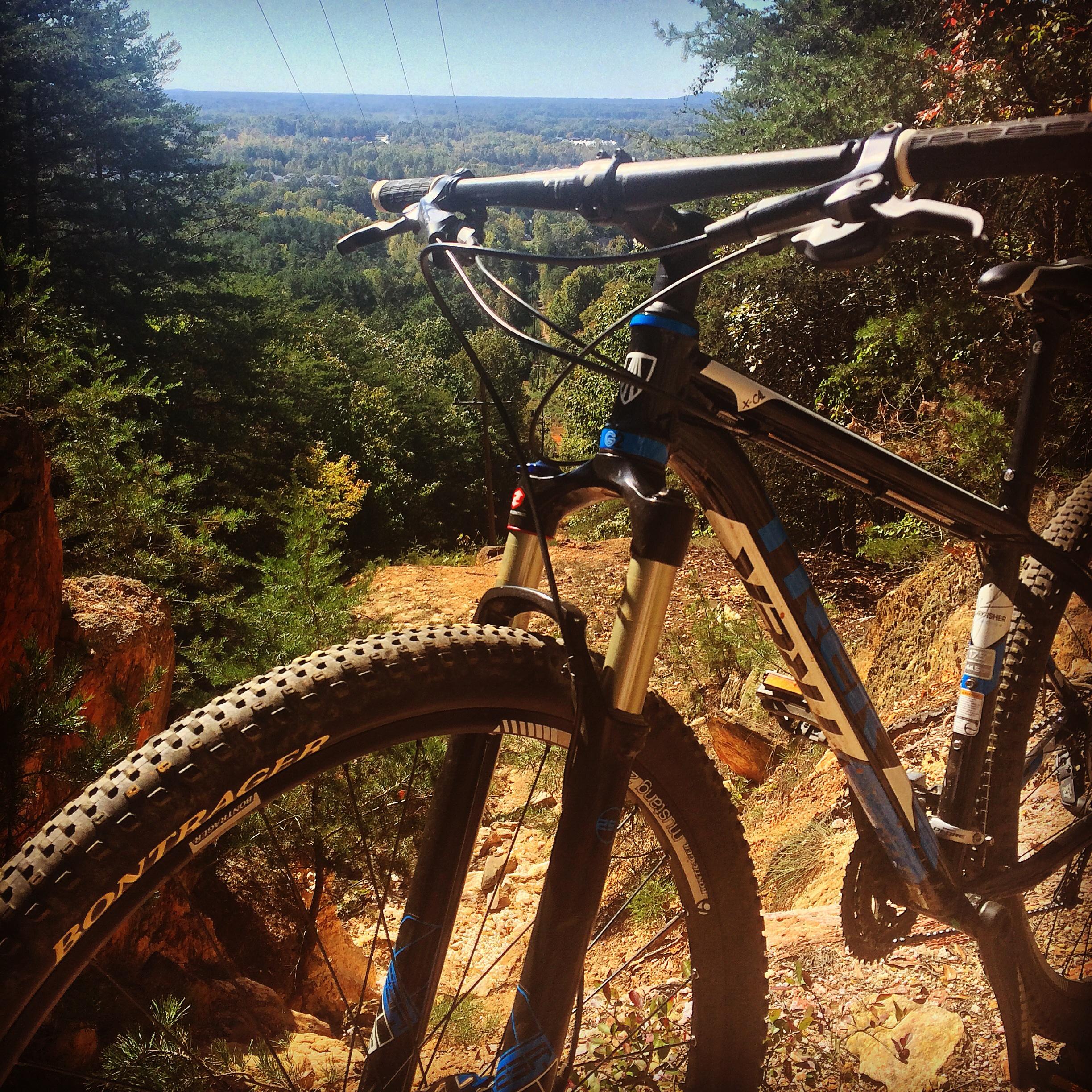 Trek X-Caliber: A close-up view of a mountain bike resting on rocky terrain, with a scenic backdrop of lush green trees and rolling hills. The handlebars and front wheel of the bike are visible, and the landscape stretches into the distance under a clear blue sky.