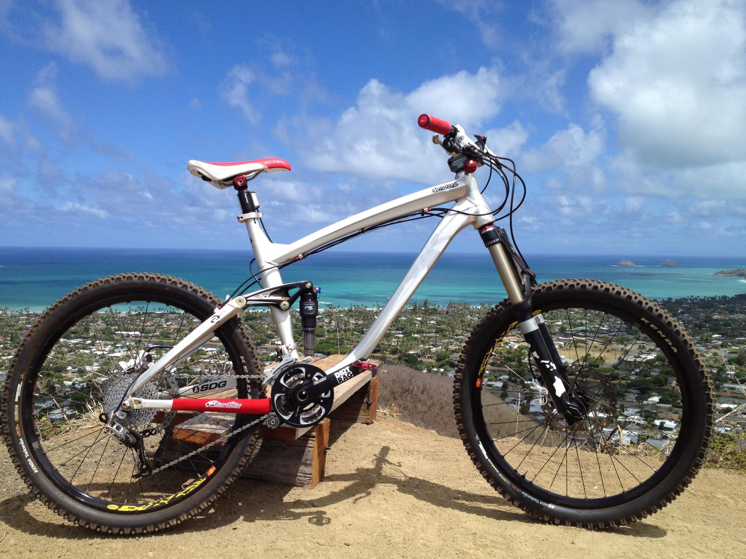 Trek Remedy 9: Mountain bike positioned on a rocky outcrop, overlooking a picturesque coastal landscape with turquoise waters, distant islands, and a clear blue sky dotted with clouds. The bike has a white frame, red accents, and thick tires suitable for rugged terrain.