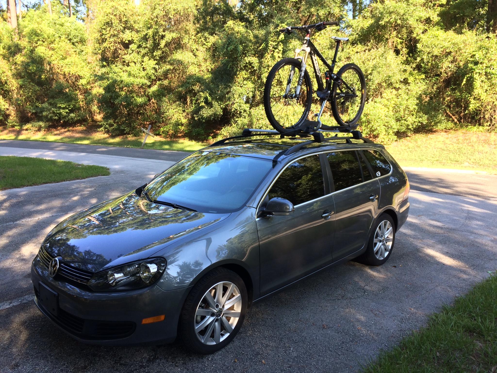 Trek Fuel EX: A gray Volkswagen car parked beside a road, featuring a mountain bike securely mounted on its roof rack. The scene is set in a natural landscape with green foliage in the background.