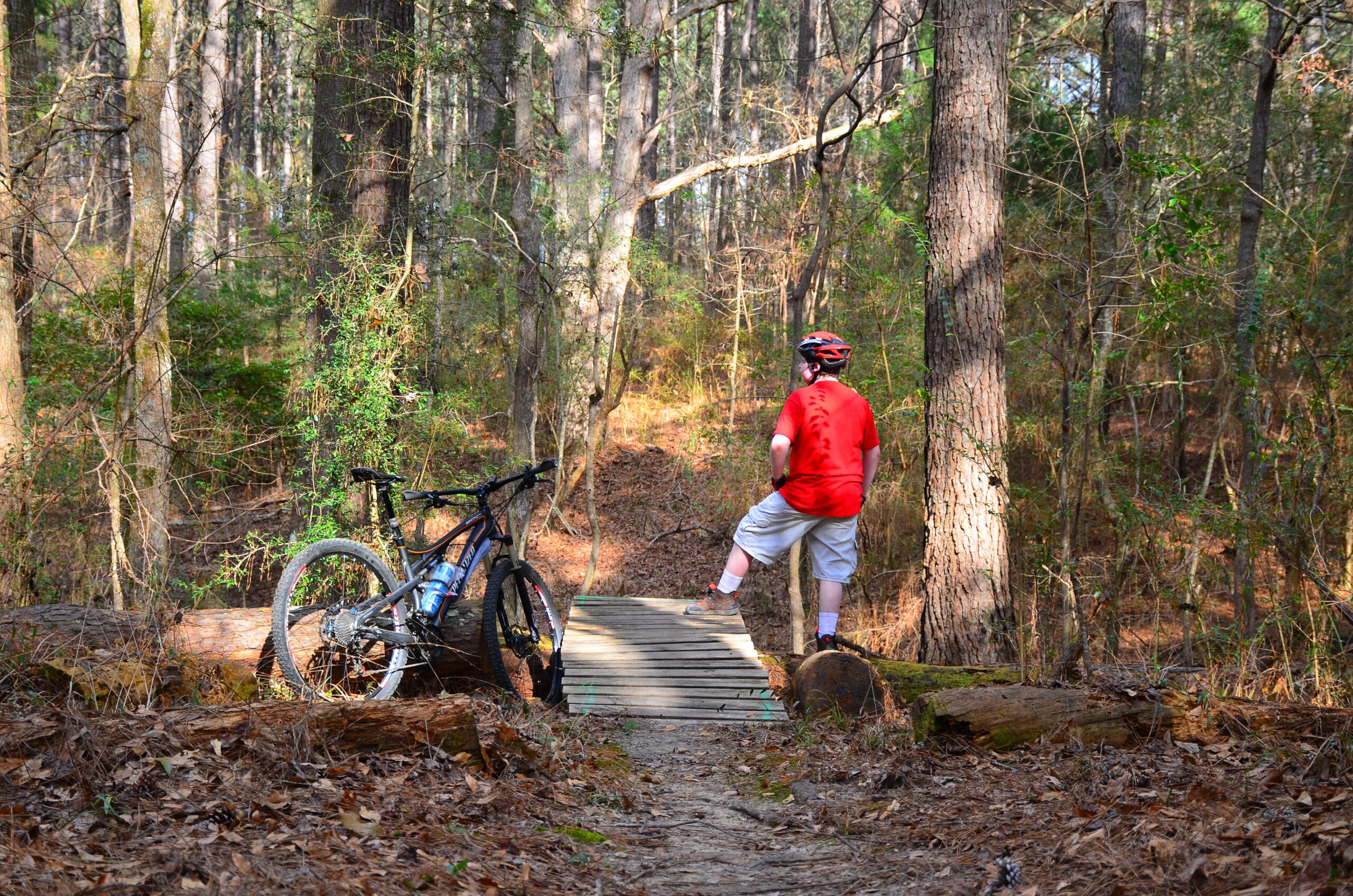 A person in a red shirt and helmet stands on a wooden bridge in a forested area, looking out into the trees. A mountain bike is propped against a log nearby, with fallen leaves and branches scattered on the ground. The scene captures a moment of contemplation in a natural setting. Mt. Zion Bike Trails mountain bike trail.