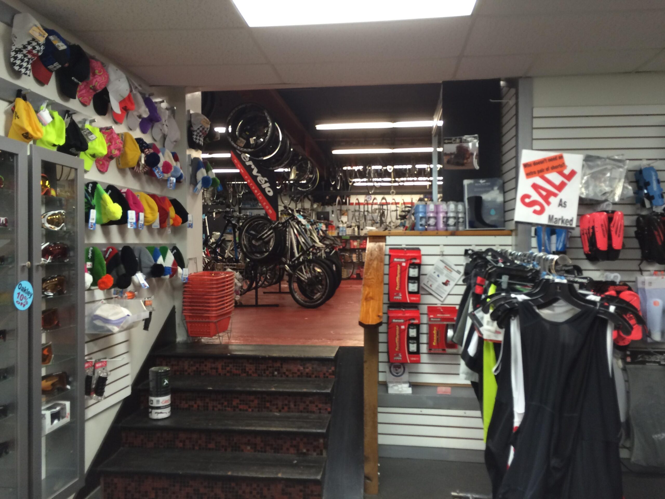 A view of a bicycle shop interior featuring a variety of cycling gear and accessories. The left wall displays colorful hats and sunglasses, while shelves on the right showcase clothing items and bike-related products. In the background, bicycles are prominently displayed, and a "SALE" sign is visible above merchandise. The shop has a vibrant and organized layout with a welcoming atmosphere.