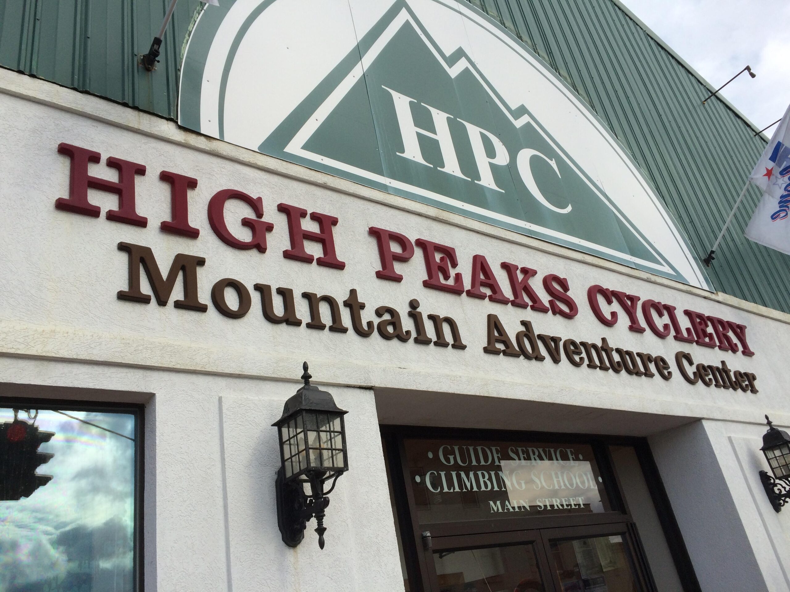 Signage of "High Peaks Cyclery Mountain Adventure Center" displayed on a building exterior, featuring large letters along with additional services like "Guide Service" and "Climbing School." There are decorative light fixtures on either side of the entrance. The background shows a green metal roof with a logo above the text.