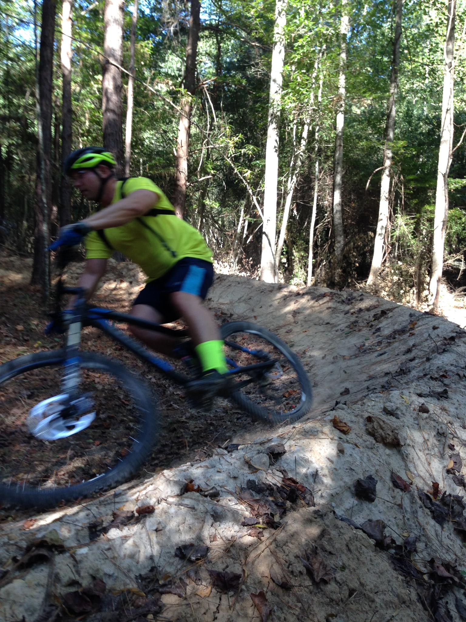A cyclist in a bright yellow shirt and green socks rides a mountain bike along a dirt trail in a wooded area, surrounded by tall trees and sunlight filtering through the leaves. The bike is leaning to the side as the rider navigates a sandy bank, demonstrating speed and agility. Mt. Zion Bike Trails mountain bike trail.