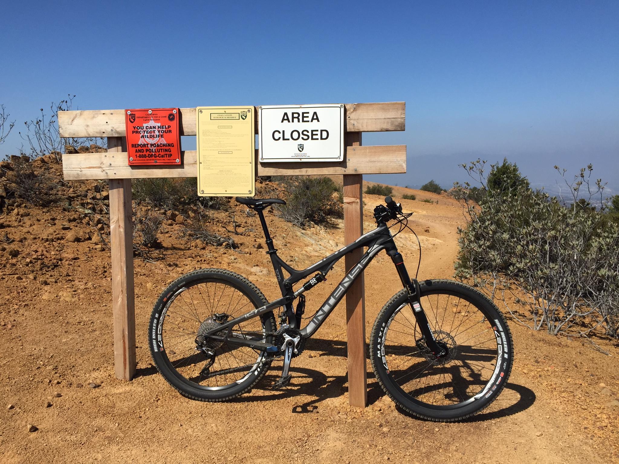Intense Tracer 275: A black mountain bike resting next to a sign marking a closed area, with additional signs about wildlife protection and reporting poaching. The background features a clear blue sky and a dirt trail leading into the distance, surrounded by sparse vegetation.