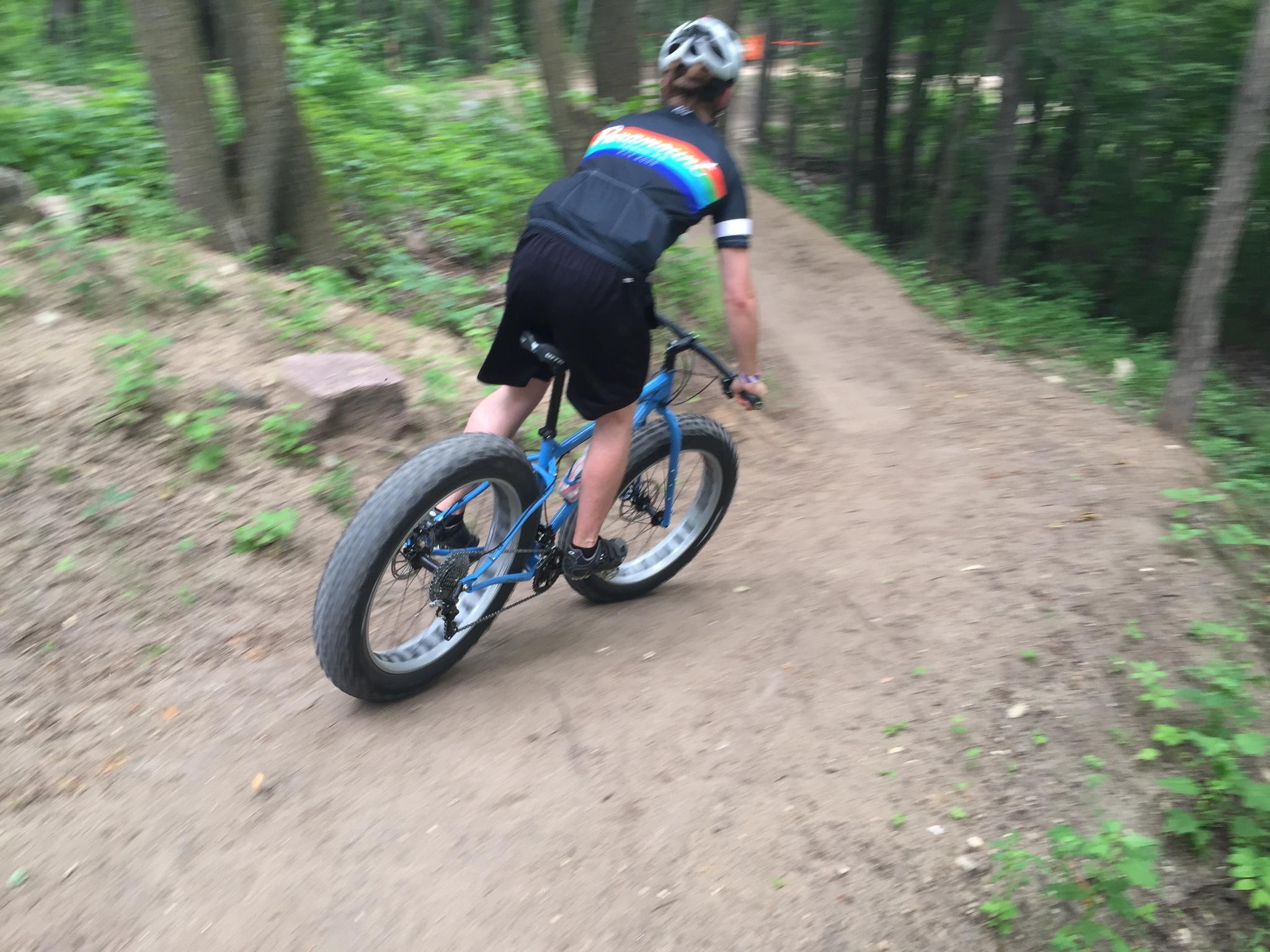Surly Ice Cream Truck: A cyclist riding a blue fat bike on a dirt trail surrounded by trees and greenery, captured in motion, with the focus on the bike's large tire and the rider's posture.