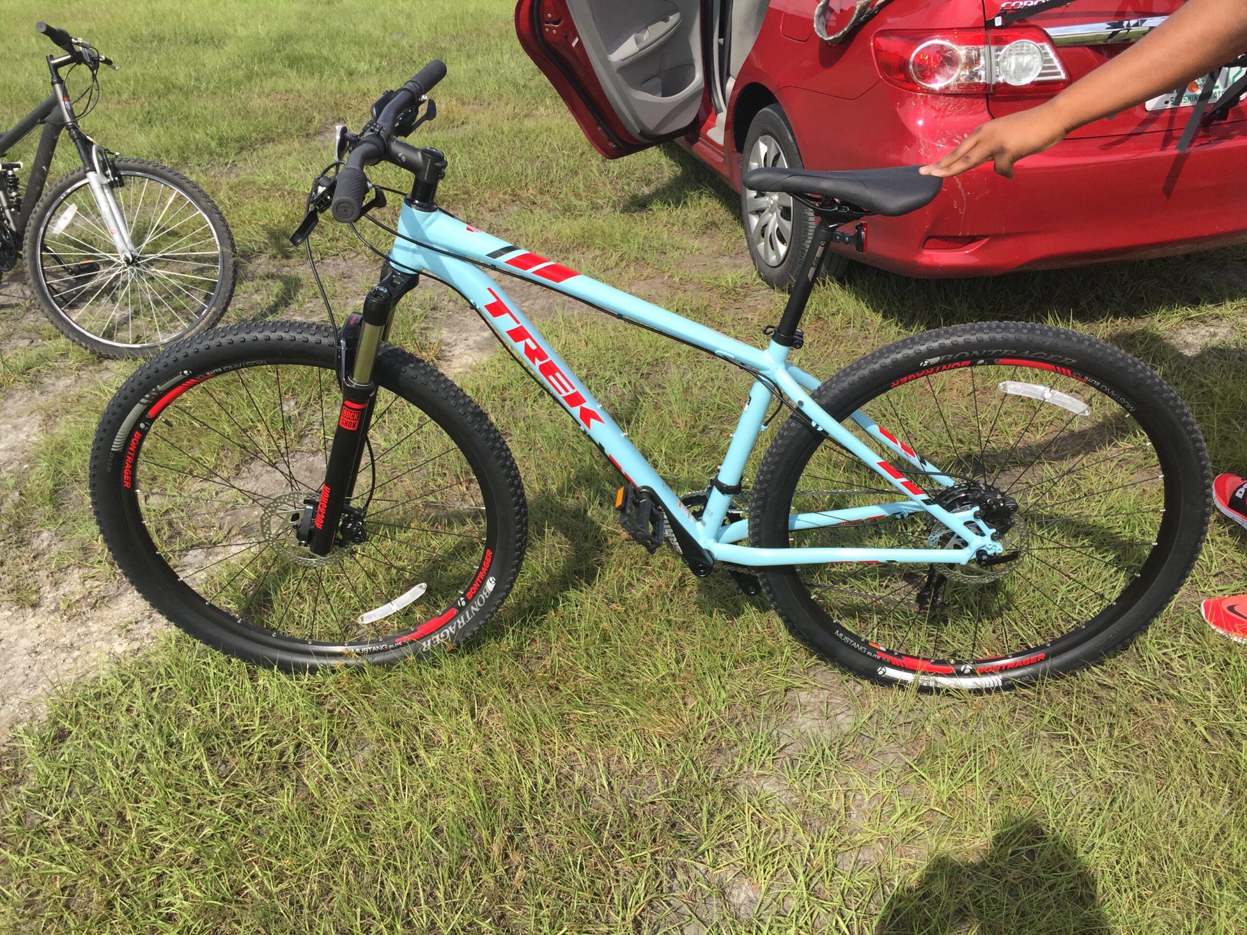 Trek EX 8: A blue Trek mountain bike parked on grass, with a black bike in the background. A person's hand is touching the bike seat, and a red car is visible in the background.