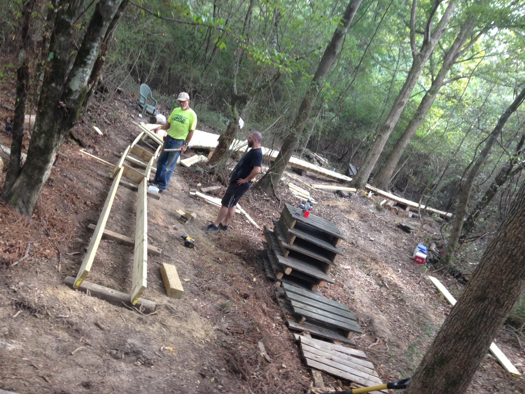 Two men are working in a wooded area, discussing the construction of a wooden structure. One man wears a bright green shirt and a hat, while the other is in a black shirt. They are surrounded by various wooden planks and pallets scattered on the ground, with trees and greenery in the background. A chair can be seen in the distance, and tools are placed nearby. Mt. Zion Bike Trails mountain bike trail.