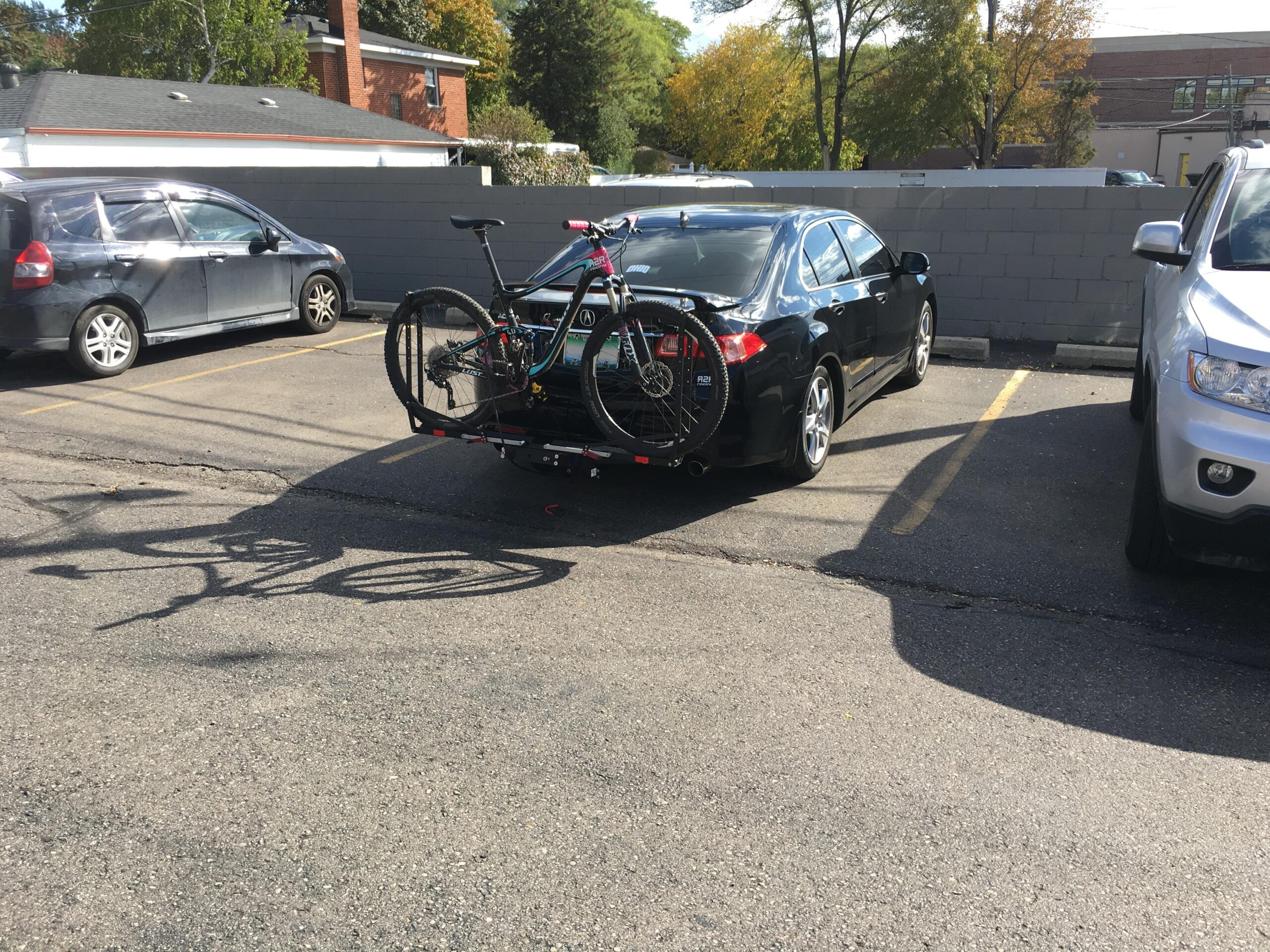 Giant Lust Advanced 2: A black car parked in a lot with two bicycles mounted on the rear bike rack. The car is surrounded by other vehicles and there is a gray wall in the background. The asphalt shows shadows cast by the bikes and the cars nearby, with some trees visible in the background indicating a sunny day.