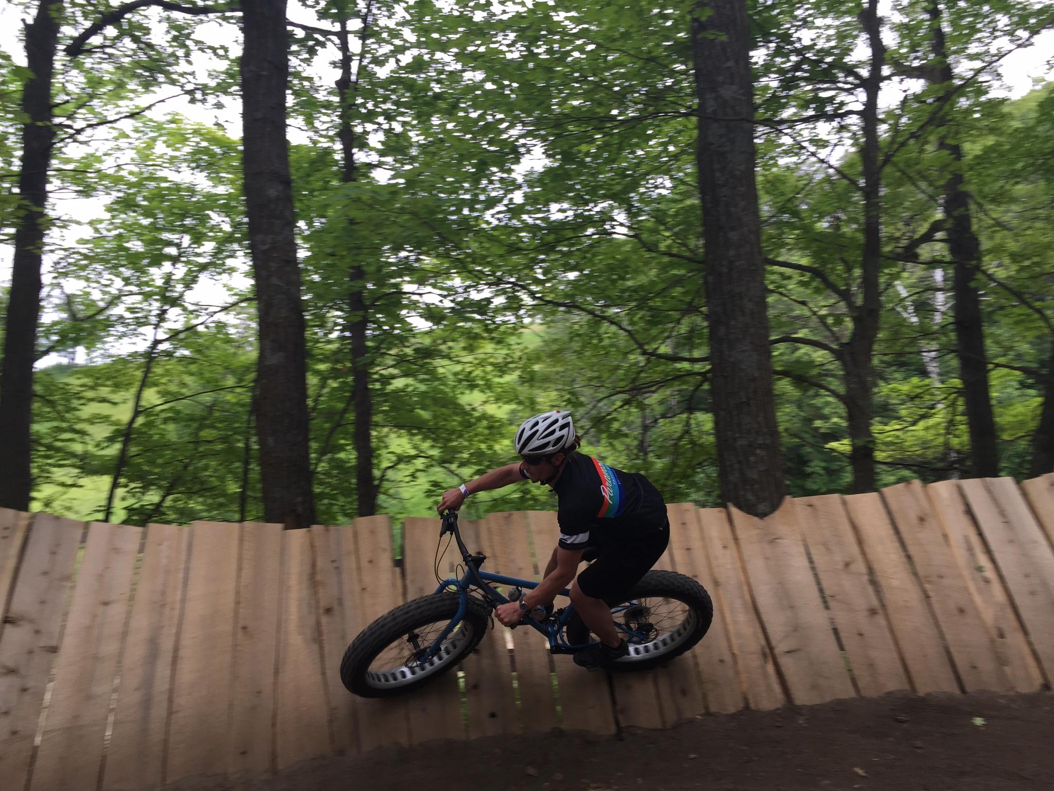 Surly Ice Cream Truck: A mountain biker maneuvering along a wooden ramp in a forested area, surrounded by lush green trees. The rider is leaning into the turn, showcasing skill and agility on the trail.