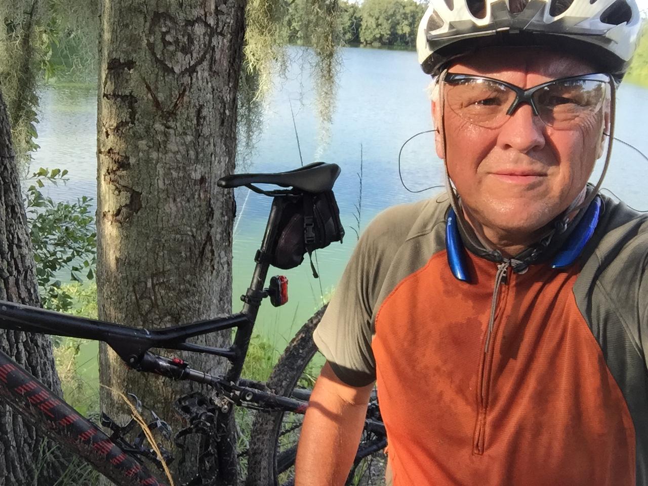 A person wearing a cycling helmet and sunglasses poses for a selfie next to a mountain bike parked against a tree. The background features a serene body of water surrounded by greenery, with some moss hanging from the tree branches. The individual appears to have just finished a ride, as they are wearing a sweat-dampened cycling jersey. Loyce E. Harpe Park mountain bike trail.