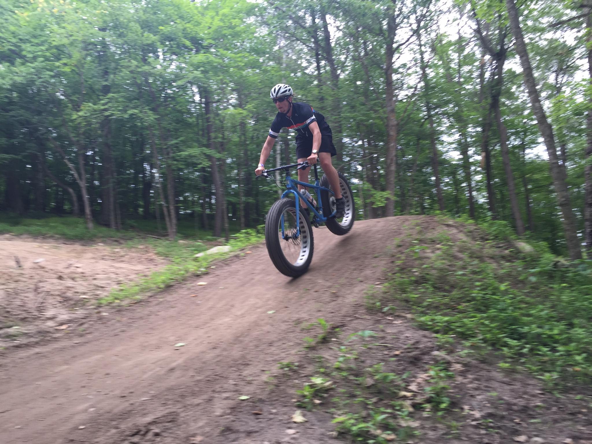 Surly Ice Cream Truck: A person riding a fat bike, displaying a jump on a dirt ramp in a forested area, surrounded by green trees and foliage. The rider is wearing a helmet and athletic clothing, capturing an action shot in mid-air while navigating the terrain.