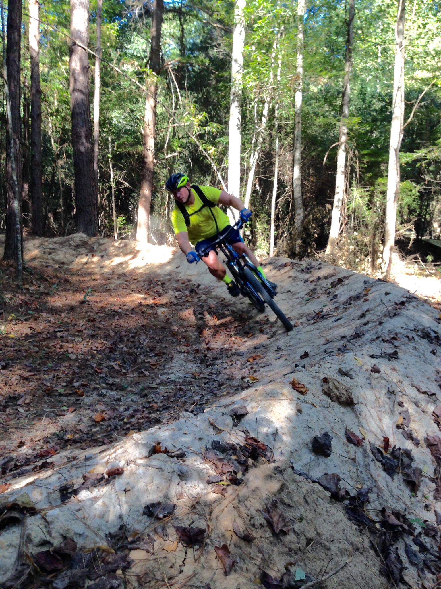 A mountain biker riding along a dirt trail in a forested area, surrounded by trees and scattered autumn leaves. The biker is leaning into a turn, wearing a bright yellow shirt, helmet, and gloves. Sunlight filters through the trees, highlighting the trail. Mt. Zion Bike Trails mountain bike trail.