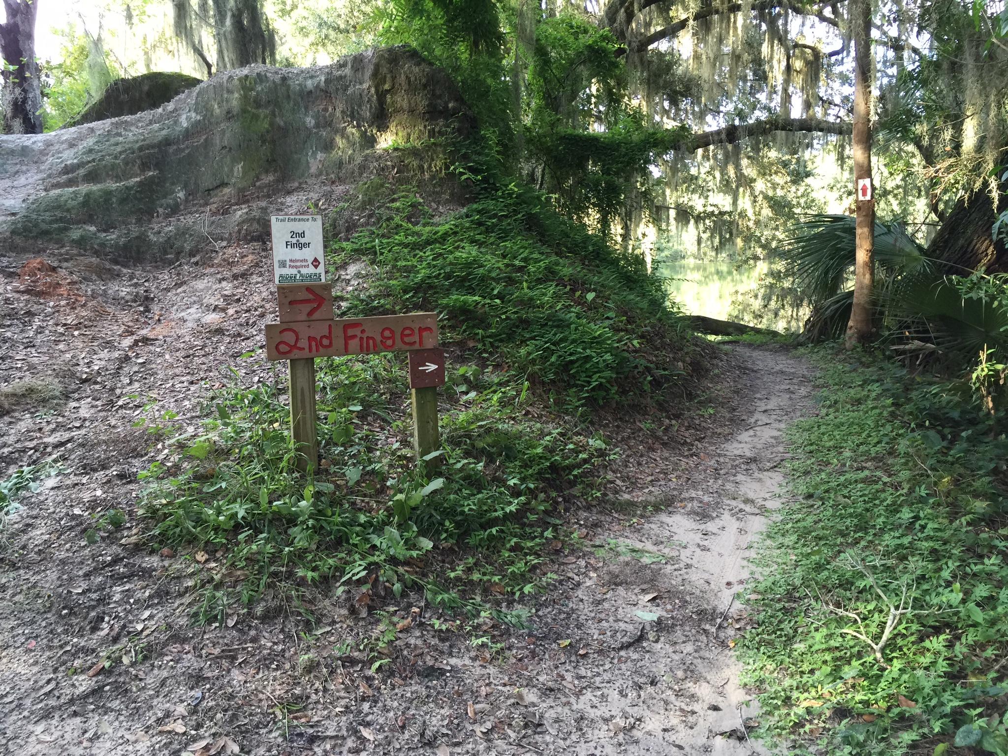 A trail sign labeled "2nd Finger" points towards a sandy path leading through a lush green area. The surrounding landscape features dense foliage, trees, and moss hanging from branches, indicating a natural outdoor environment. A small sign indicating the trail entrance is visible at the top of the path. Loyce E. Harpe Park mountain bike trail.