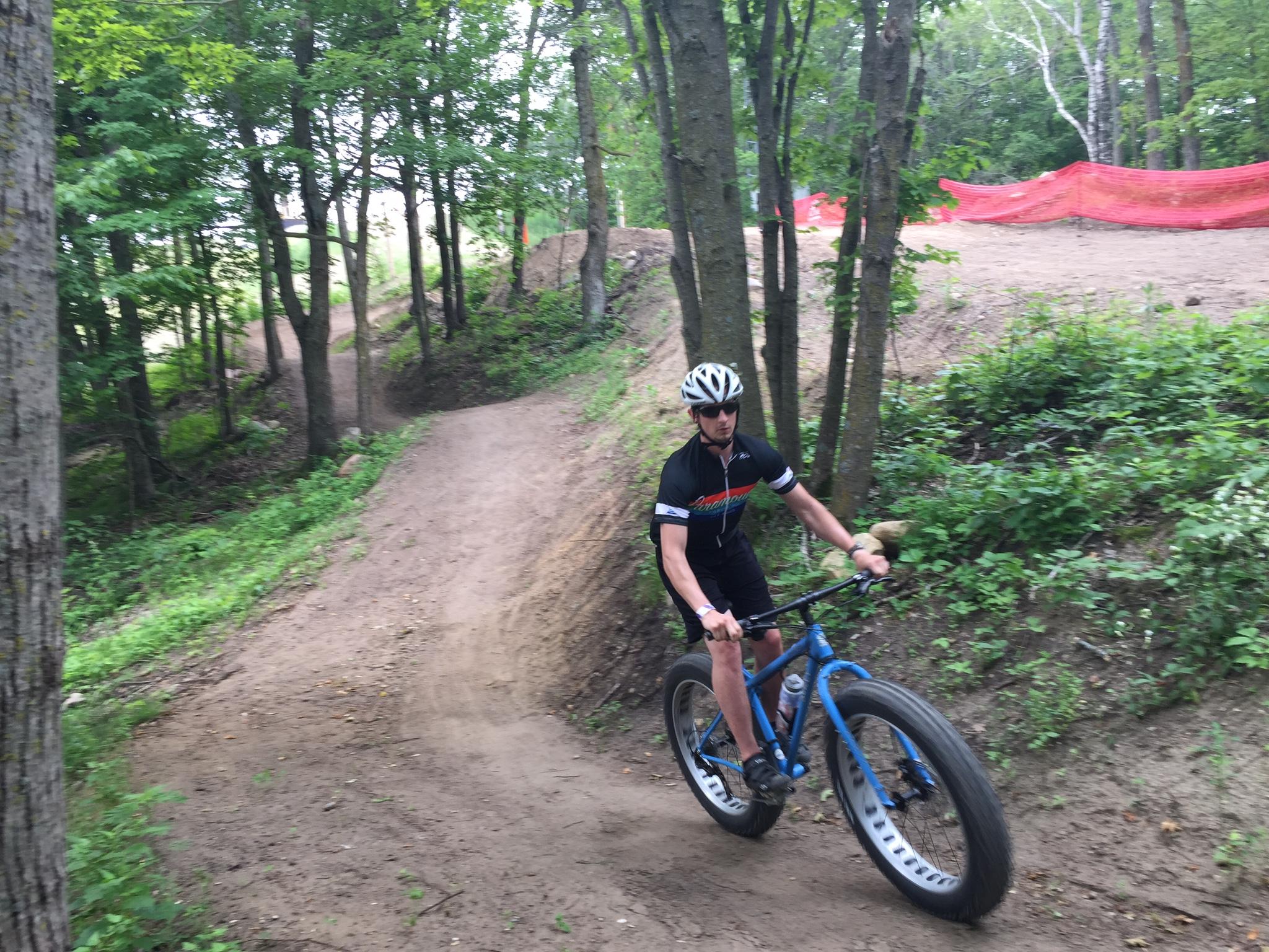 Surly Ice Cream Truck: A cyclist rides a blue fat bike along a twisty dirt trail in a forested area, surrounded by trees and greenery. The path leads uphill, with a sandy texture, and a barrier can be seen in the background. The cyclist is wearing a helmet and a black cycling jersey.