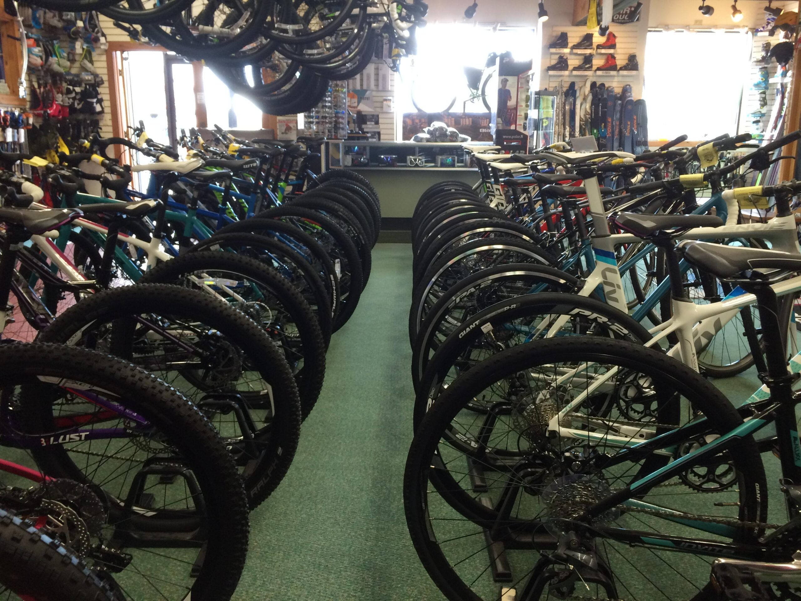 A bike shop interior featuring a row of mountain bikes on display. The scene includes various colored bicycles with their wheels prominently shown, while additional bikes are visible in the background. The shop is well-lit, showcasing bicycle accessories and gear on the walls.
