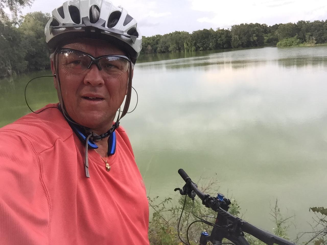 A person wearing a bicycle helmet and sunglasses takes a selfie near a calm lake surrounded by trees. They are dressed in a bright orange cycling shirt and have a mountain bike resting beside them. The overcast sky reflects softly on the water's surface. Loyce E. Harpe Park mountain bike trail.