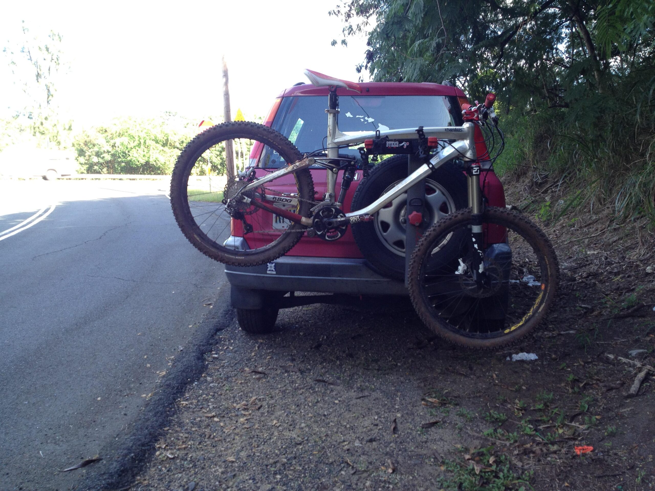 Trek Remedy 9: A red vehicle parked on the side of a road, with a mountain bike mounted on the rear. The bike, which has a dirty frame and tires, is secured to a bike rack. Surrounding the vehicle are patches of grass and trees, with a glimpse of another car visible in the background.
