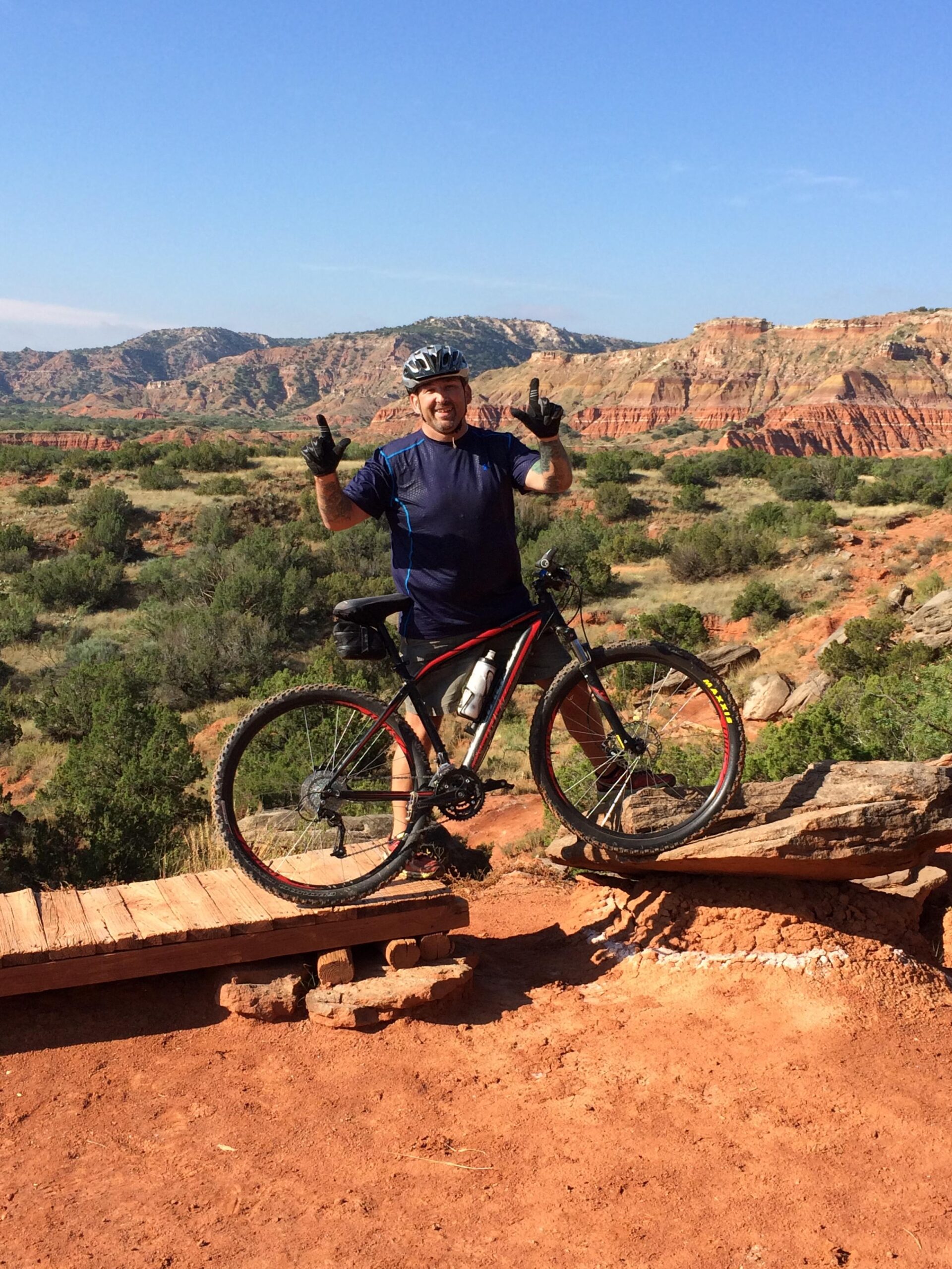 Specialized Rockhopper Sport 29: A man in a blue athletic shirt and gloves stands on a mountain bike, posing with a joyful expression and making a hand gesture. He is on a rocky path with a scenic backdrop of red rock formations and green shrubs under a clear blue sky.