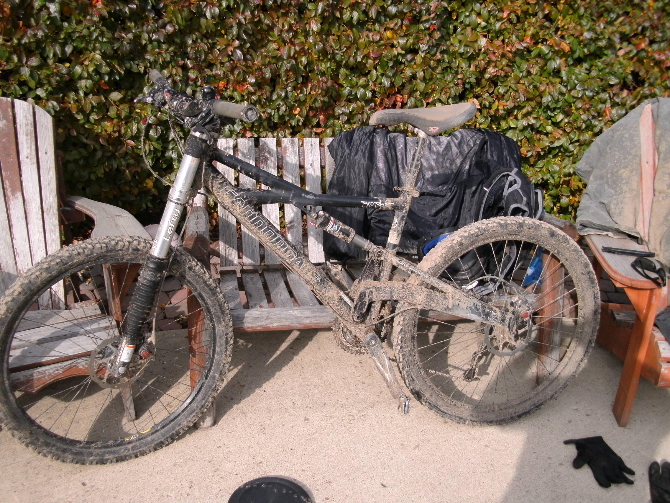 Cannondale Prophet: A muddy mountain bike parked on a wooden bench, with a black bag in the background and a pair of gloves on the ground. The bike shows signs of recent use, with dirt covering the frame and tires.