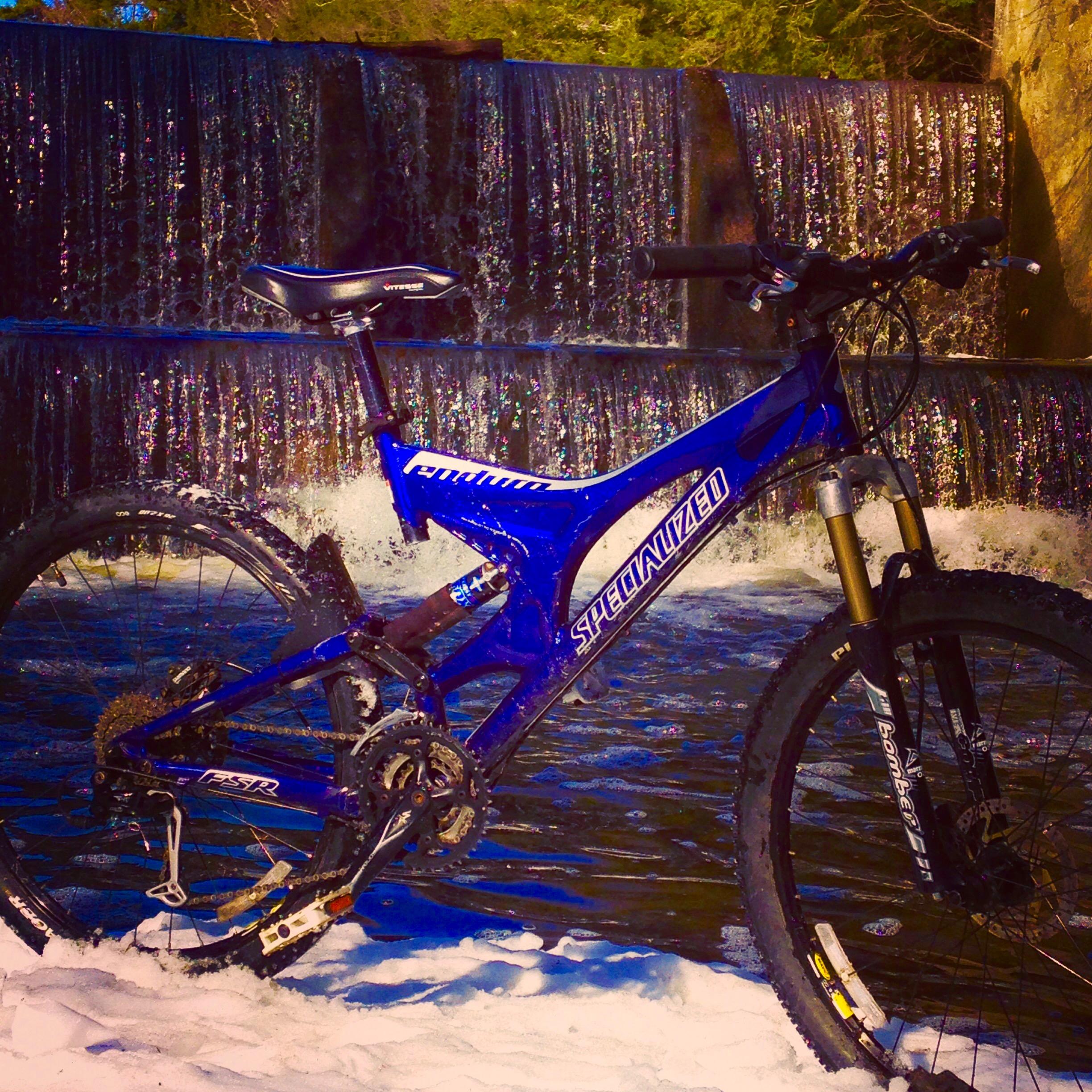 A blue mountain bike resting on snow in front of a cascading waterfall, with sunlight reflecting off the water. The bike features a sturdy frame and thick tires, highlighting its suitability for outdoor adventures.