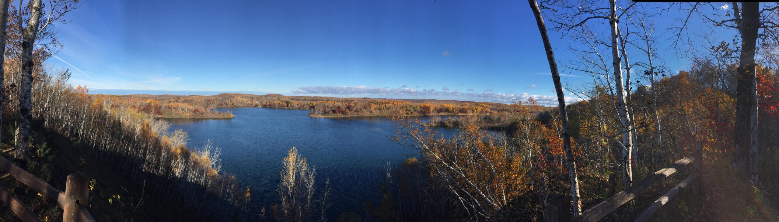 A panoramic view of a serene lake surrounded by autumn foliage. The landscape features vibrant orange, yellow, and red leaves on trees, contrasting against a clear blue sky. The calm water reflects the colorful trees and the distant hills, while a wooden fence lines the foreground, enhancing the scenic beauty of the natural setting. Cuyuna Lakes mountain bike trail.
