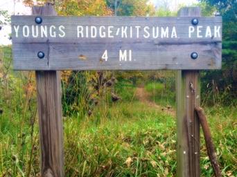 Wooden sign marking the Youngs Ridge/Kitsuma Peak hiking trail, indicating a distance of 4 miles. The background features tall grass and trees, suggesting a natural setting. Kitsuma mountain bike trail.
