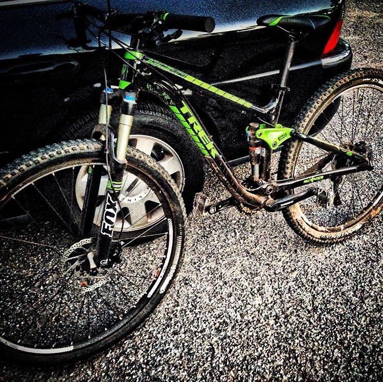 Trek Fuel EX 7 29: A close-up view of a black mountain bike with green accents, parked next to a car. The bike features thick, knobby tires and front suspension forks, indicating it's designed for off-road riding. The surrounding area is gravel, suggesting a past ride on rugged terrain.