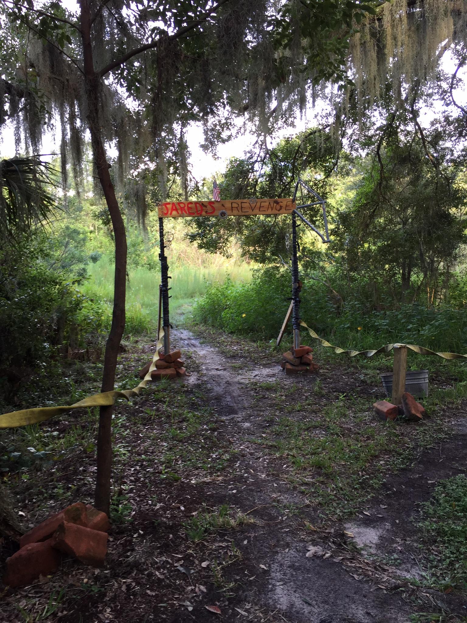 A wooded pathway leading into a lush green area, marked by a wooden sign that reads "JARED'S REVENGE." The entrance is framed by two posts, with yellow caution tape and orange brick supports visible on the ground. Spanish moss hangs from the trees overhead, creating a natural, shaded atmosphere. Loyce E. Harpe Park mountain bike trail.