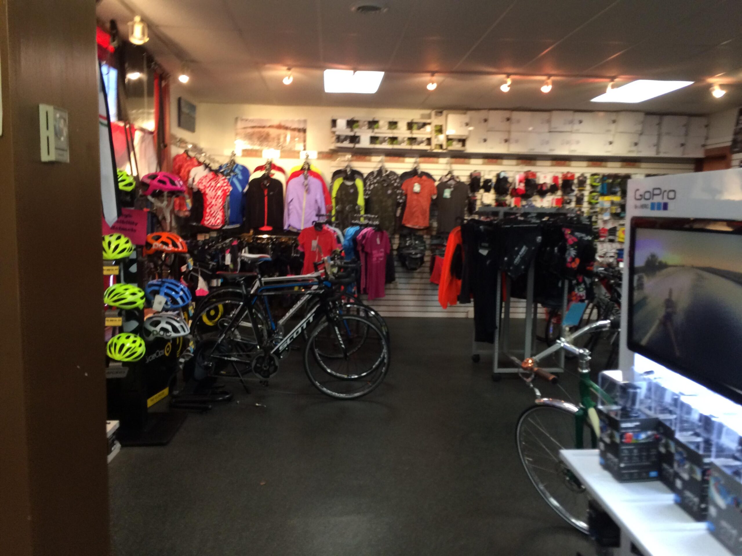 Interior view of a sports shop showcasing bicycles, various color helmets, cycling apparel, and accessories. The store features racks of clothing in bright colors, with items displayed on walls and tables. A GoPro display can be seen in the foreground. The atmosphere is vibrant and geared towards cycling enthusiasts.