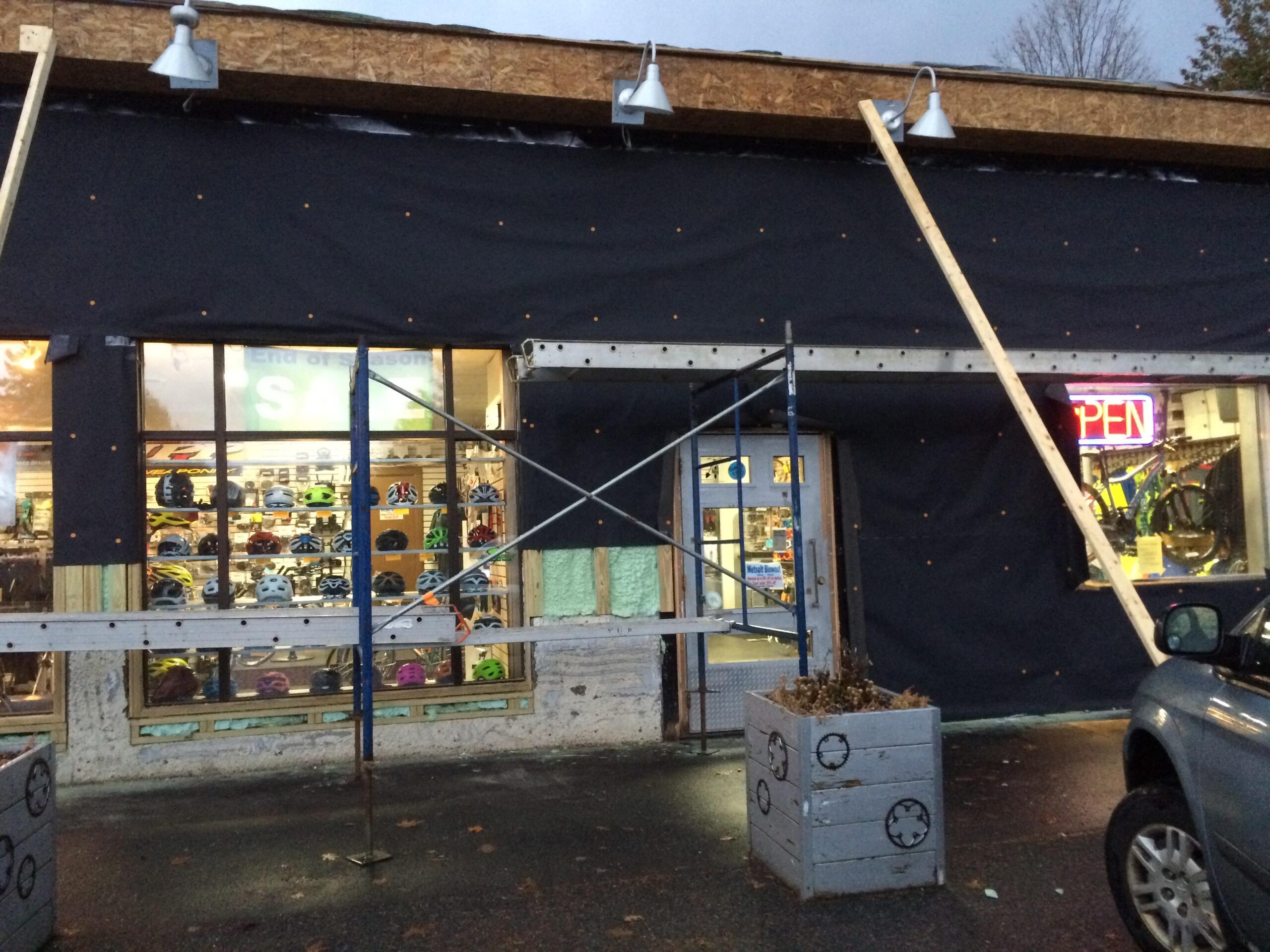 Image of a storefront under renovation, featuring a black tarp covering part of the exterior. Scaffolding is visible in front of the entrance, with construction lights mounted above. Through the windows, displays of colorful helmets can be seen, along with an illuminated "OPEN" sign. A parked vehicle is located to the right, and a planter is placed in front of the building.