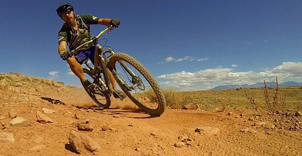 A mountain biker navigating a dusty dirt trail, leaning into a turn on a sunny day with a blue sky and distant mountains in the background. The scene captures the action and excitement of off-road cycling. Moab Brand Trails mountain bike trail.