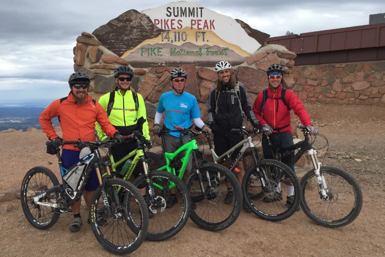 Five mountain bikers pose with their bikes in front of the summit sign for Pikes Peak, which reads "Summit Pikes Peak 14,110 ft. Pike National Forest." The group is wearing various cycling gear, including helmets and brightly colored clothing. The background features a scenic view of the landscape beneath a cloudy sky.