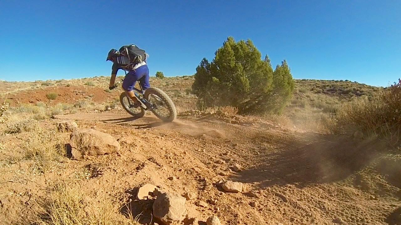 A person riding a fat tire bike on a dirt trail, kicking up dust while maneuvering around rocks and vegetation in a rugged outdoor landscape under a clear blue sky. Moab Brand Trails mountain bike trail.