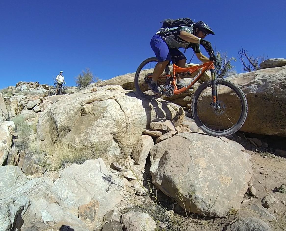 A mountain biker navigates over rocky terrain, skillfully balancing on an orange bicycle. In the background, another cyclist watches from a distance, surrounded by a desert landscape under a clear blue sky. Moab Brand Trails mountain bike trail.