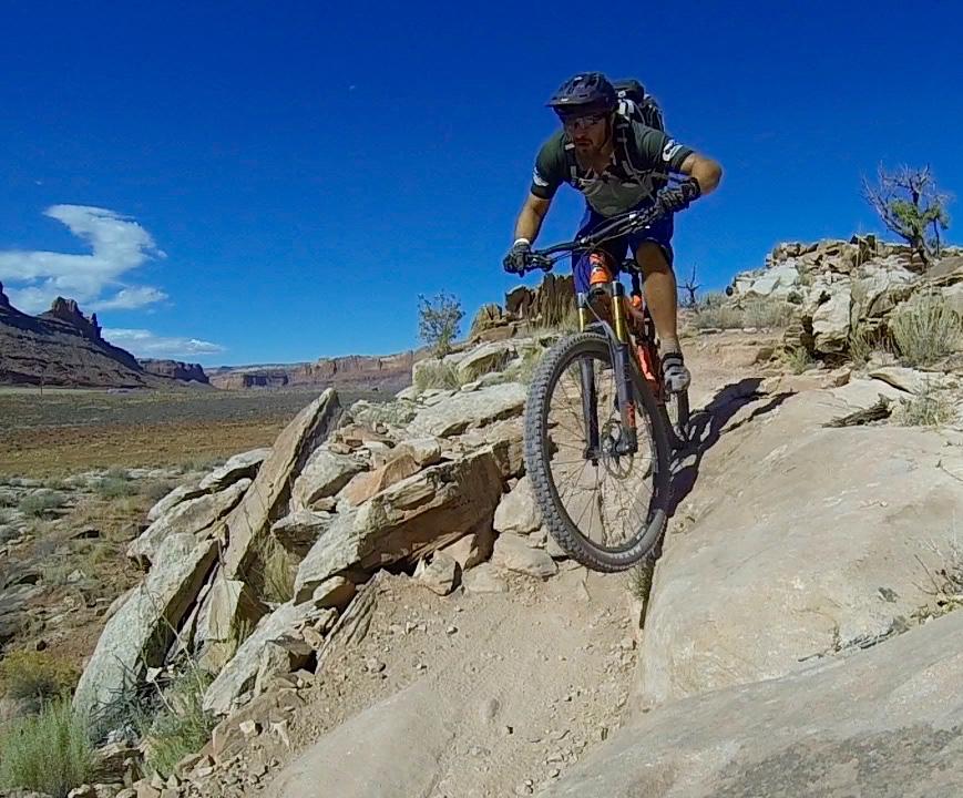 A mountain biker navigating a rocky trail with a steep incline, surrounded by a desert landscape and blue sky. The biker is wearing a helmet and has a focused expression as they maneuver over the uneven terrain. Moab Brand Trails mountain bike trail.