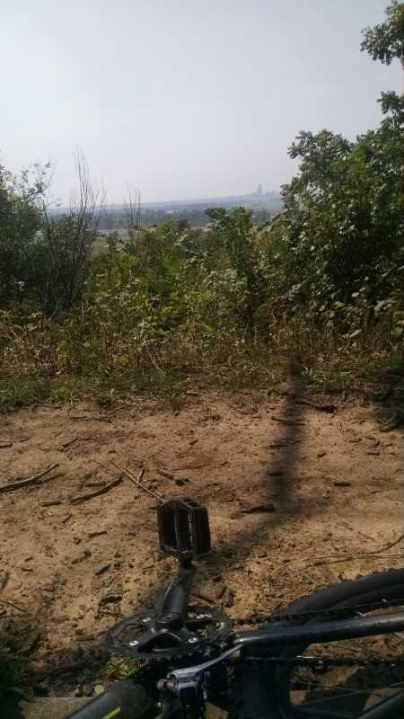 A scenic view from a hiking trail overlooking a valley, with green foliage and shrubs in the foreground. A bicycle pedal is positioned at the bottom of the image, suggesting a biking adventure. The sky is hazy, indicating a warm, possibly sunny day. Lewis And Clark Monument mountain bike trail.