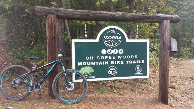 Sign for Chicopee Woods Mountain Bike Trails, featuring the IMBA logo, with a mountain bike leaning against the wooden frame. The surrounding area is wooded, indicating a natural setting for outdoor activities. Chicopee Woods mountain bike trail.