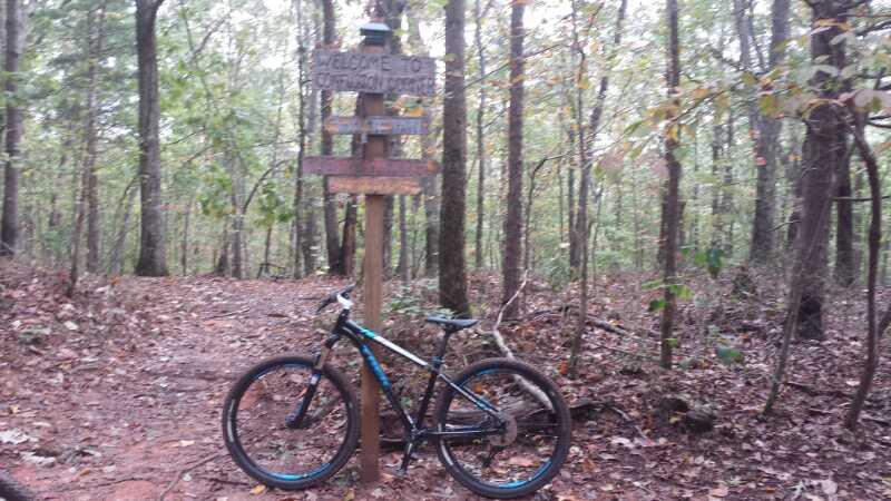 A mountain bike parked beside a wooden signpost that reads "Welcome to..." in a wooded area. The ground is covered with fallen leaves, and trees surround the path that leads deeper into the forest. Chicopee Woods mountain bike trail.