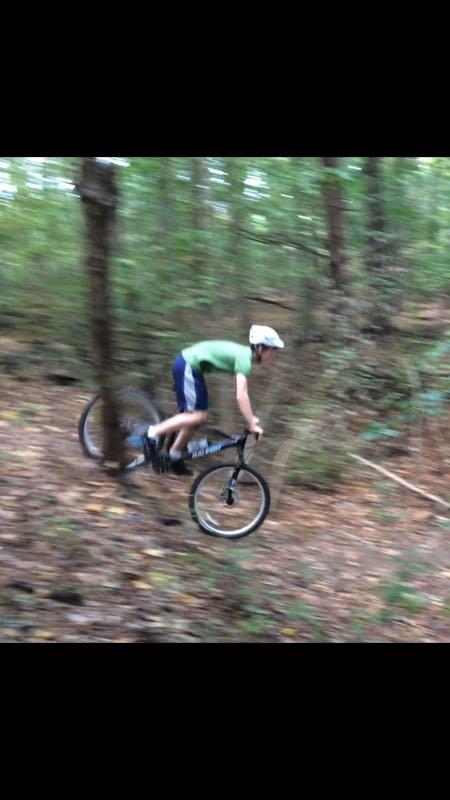 A person riding a mountain bike downhill on a wooded trail, wearing a helmet and athletic clothing. The motion blur suggests high speed as they navigate the terrain surrounded by trees and foliage. Tanglewood Park mountain bike trail.